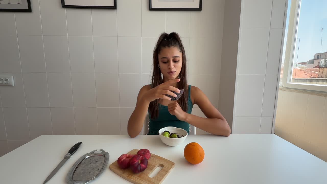 Mujer joven comiendo fruta fresca y aceitunas en una mesa de cocina