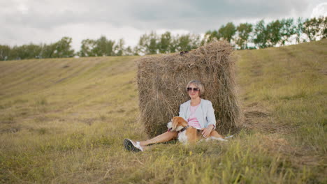 woman wearing sunglasses and casual outfit sits on dry grassy field, leaning against hay bale, she gazes into distance with a relaxed expression, surrounded by open farmland