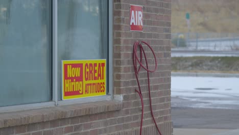 Now-hiring-great-attitudes-bright-yellow-and-red-sign-in-window-of-car-wash-with-condensation-dew-on-glass,-and-air-hose-hung-on-brick-wall-static-camera-angle-in-Cinema-4K-30fps-slowed-from-60fps