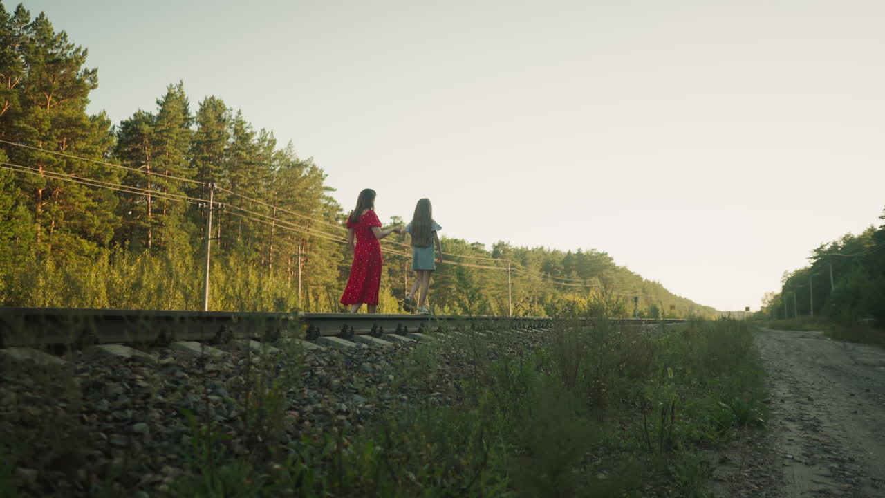 side view of woman in red dress walking alongside young girl on rail track surrounded by forest trees in warm evening light, depicting peaceful family bond and natural countryside journey