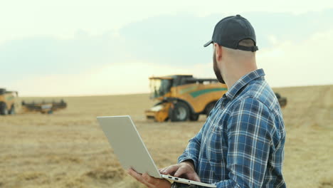 agricultor de sucesso trabalhando no computador portátil e olhando para máquinas de colheita