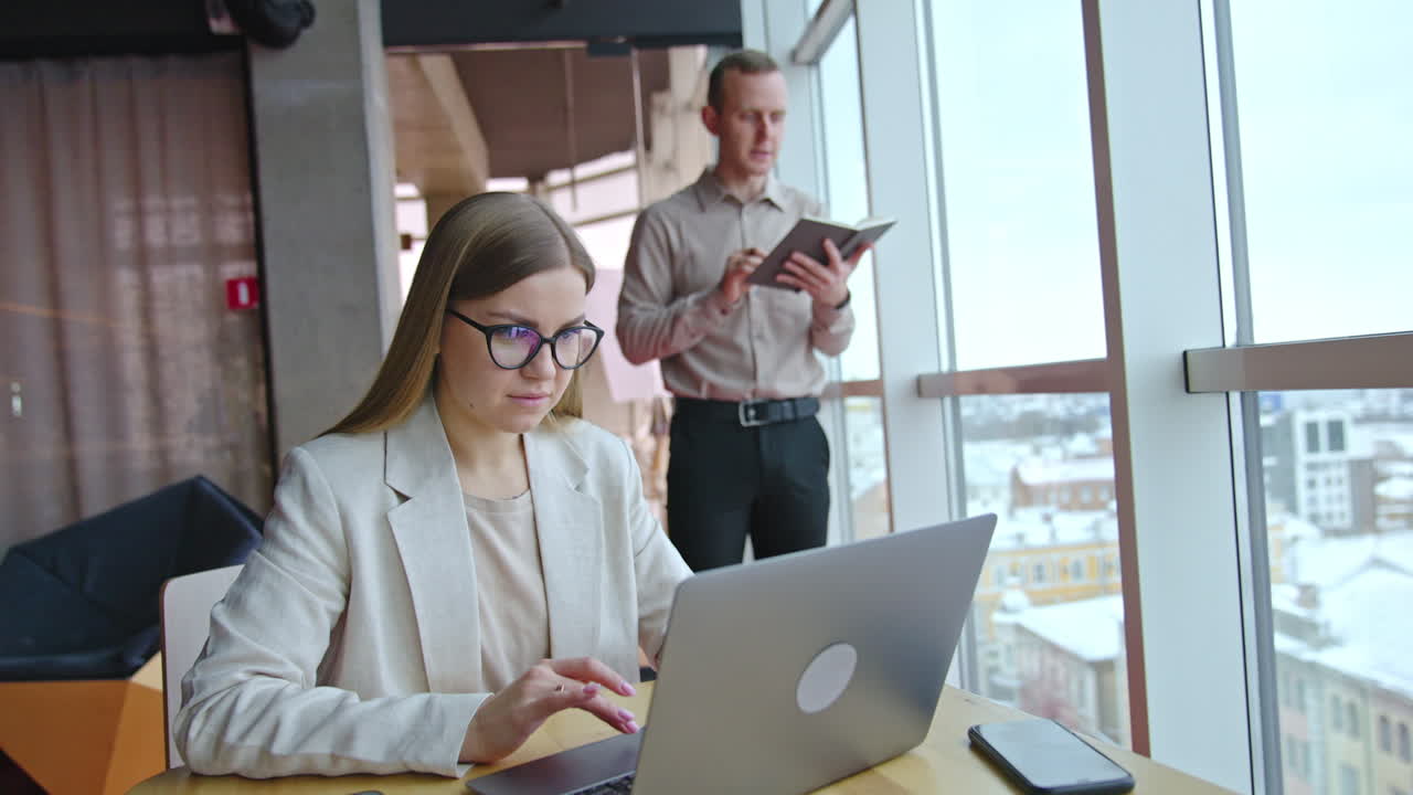 Employees working in the light office. Female typing on laptop at the desk. Male standing with notebook near the window at the backdrop.