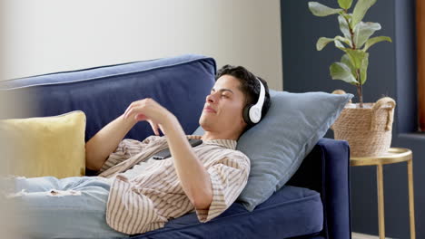 happy biracial man in headphones lying on couch using smartphone, copy space, slow motion