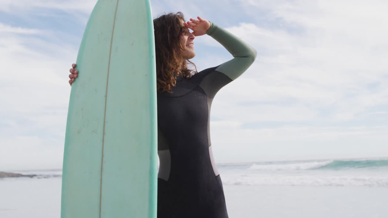 Donna felice di etnia mista in piedi su una spiaggia assolata in riva al mare con una tavola da surf, mentre guarda altrove e sorride.