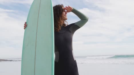 Glückliche-Frau-Mit-Gemischter-Abstammung,-Die-Am-Sonnigen-Strand-Am-Meer-Steht-Und-Ein-Surfbrett-Hält-Und-Lächelnd-Wegschaut