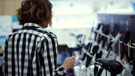 Young-cheerful-positive-woman-in-plaid-walks-choosing-curling-iron-in-household-appliances-store,-taking-one-from-the-shelf-and