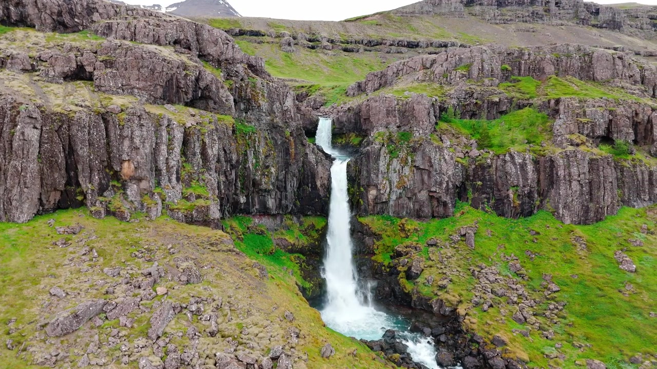 Et fantastisk luftfoto af Snadalsfoss vandfald på Island, der viser hvidt vand styrte ned ad mørke basaltklipper og grønne mosklædte bakker. Ideelt til rejse- og bæredygtighedsindhold