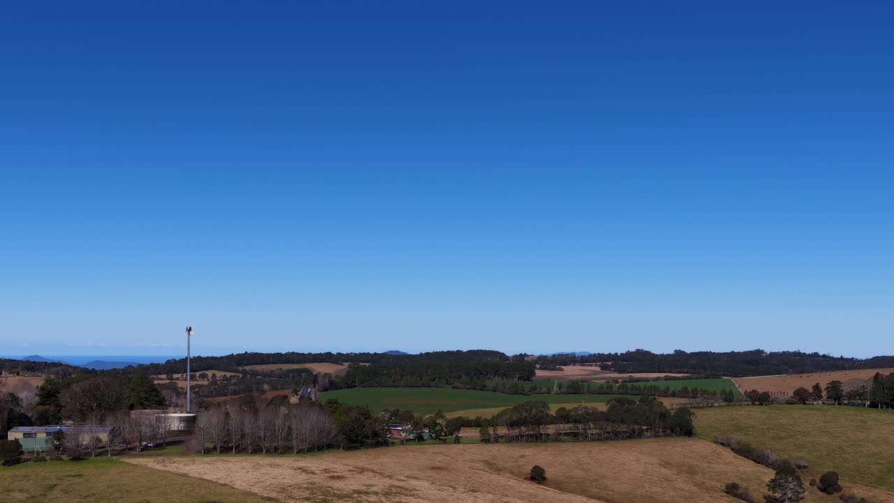 Drone ascends above sunlit rural farmland, revealing fields, trees, and distant hills under blue sky