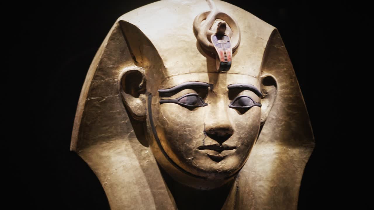 Close-up de uma máscara de caixão egípcia antiga dourada com traços faciais detalhados e ornamento de cobra, exibida sob iluminação de museu contra um fundo escuro.