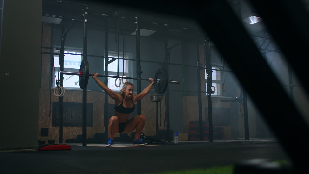 Movimento rallentato: Allenamento di squat con bilanciere sopra la testa. Donna muscolosa solleva il bilanciere sopra la testa, allenando le spalle in palestra. Atleta fitness sicura di sé pratica un allenamento di forza con attrezzature pesanti.