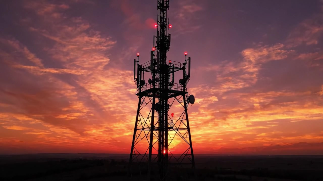 Torre di telefonia mobile in silhouette contro un cielo mozzafiato al tramonto con colori vivaci e formazioni nuvolose, che cattura l'incontro tra tecnologia moderna e bellezza naturale.