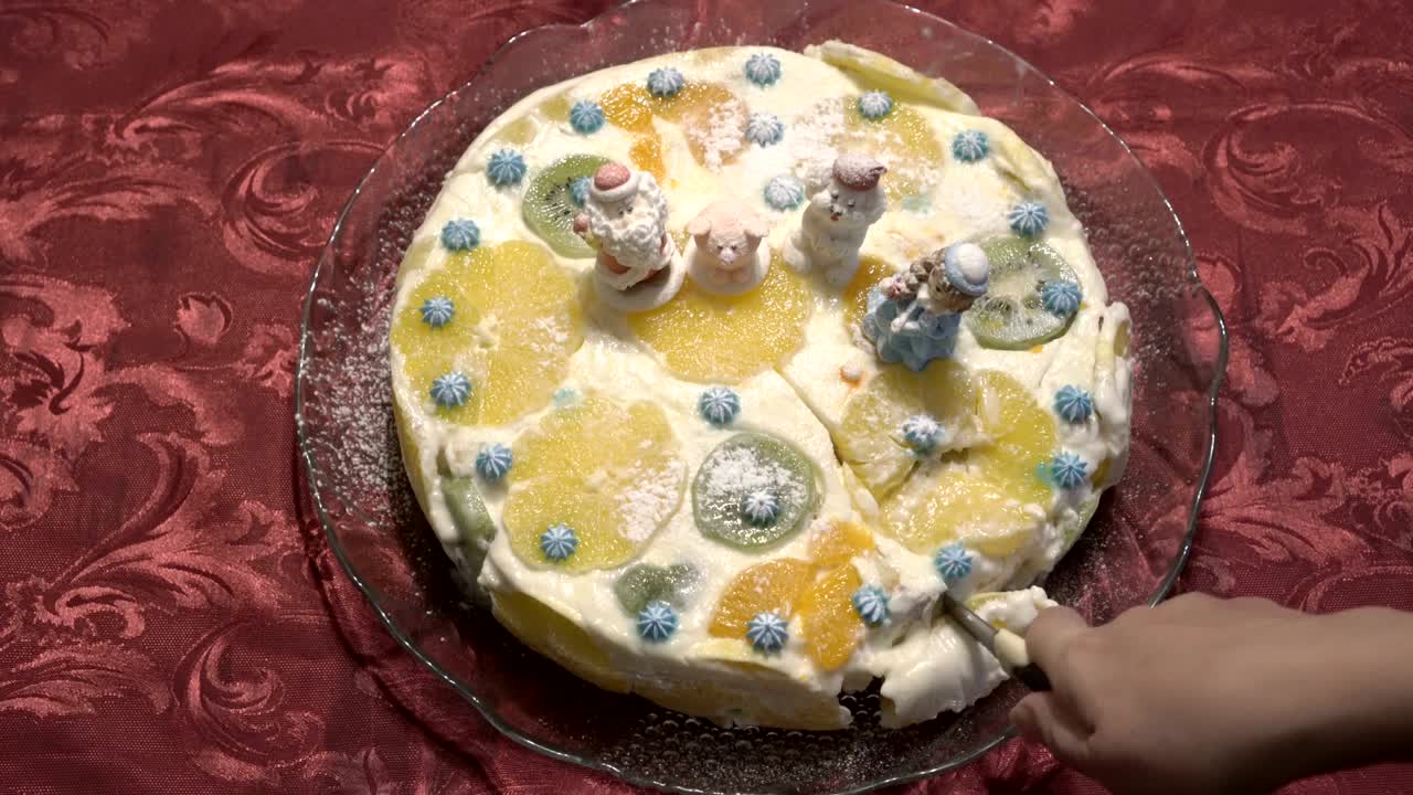 mujer corta en pedazos pastel de navidad con crema blanca y fruta, decorado con figuras de santa claus y animales. pastel vale la pena en una placa de vidrio, en manteles rojos. primer plano. la vista desde la parte superior.