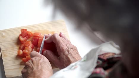 Manos-De-Una-Anciana-Cortando-Tomate-Rojo-Con-Un-Cuchillo-En-Una-Tabla-De-Corte-De-Madera,-Delantal-Blanco-Preparándose-Para-Cocinar,-Disparado-Desde-Atrás