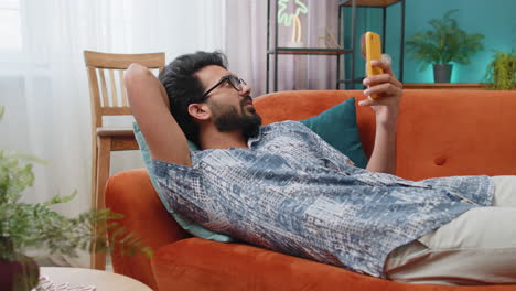 Indian-hindu-young-man-lying-on-couch,-using-smartphone-share-messages-on-social-media-application