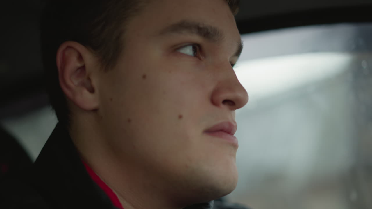 Close up young male driver focused on road ahead with determined gaze, subtle head movement in car interior with daylight through window illuminating face, wearing black jacket and seat belt strap