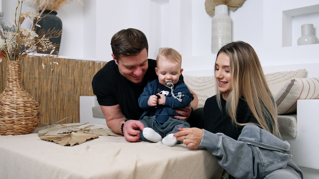 Caring parents sit on both sides from their baby. Parents tickle their child's feet and kid is playing with a bracelet.