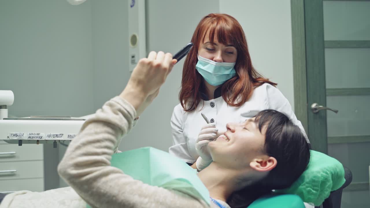 A dentist gives brunette a mirror for inspecting the result after the teeth whitening procedure at the stomatologic clinic.