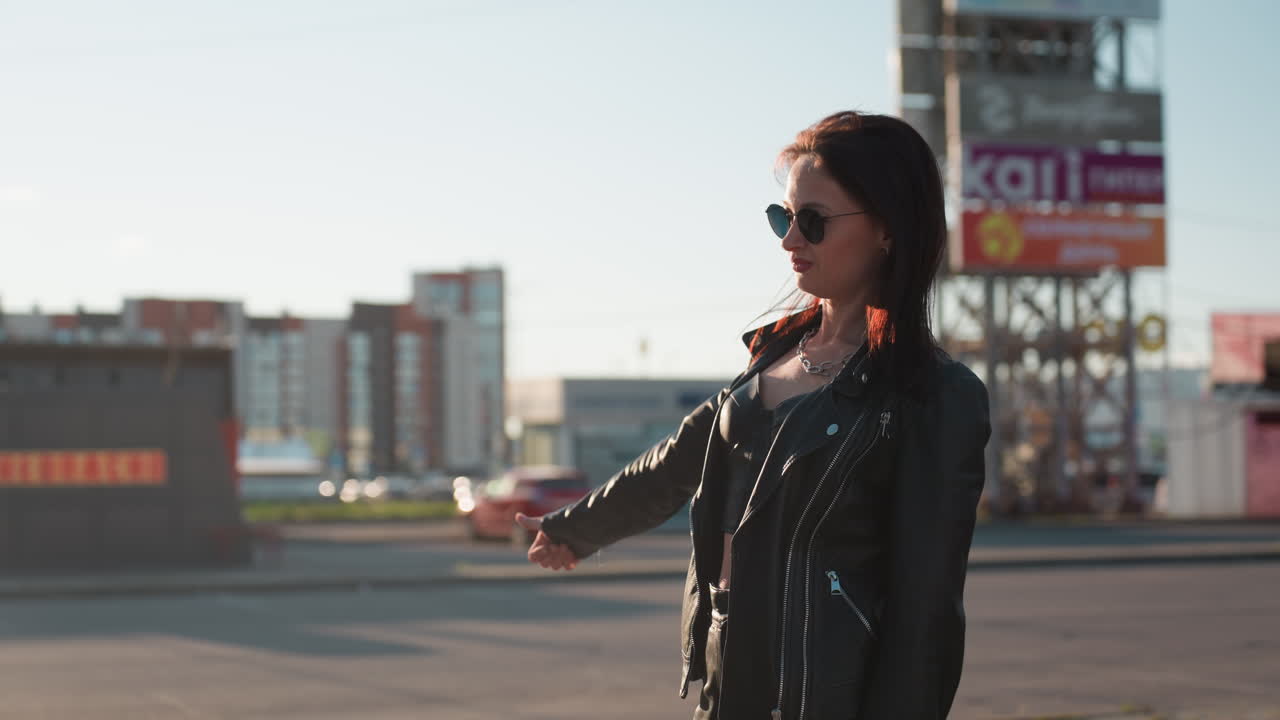 Stylish lady in black leather jacket and sunglasses stands on urban street extending her arm to signal motorcycle rider to stop before climbing and exuding confidence