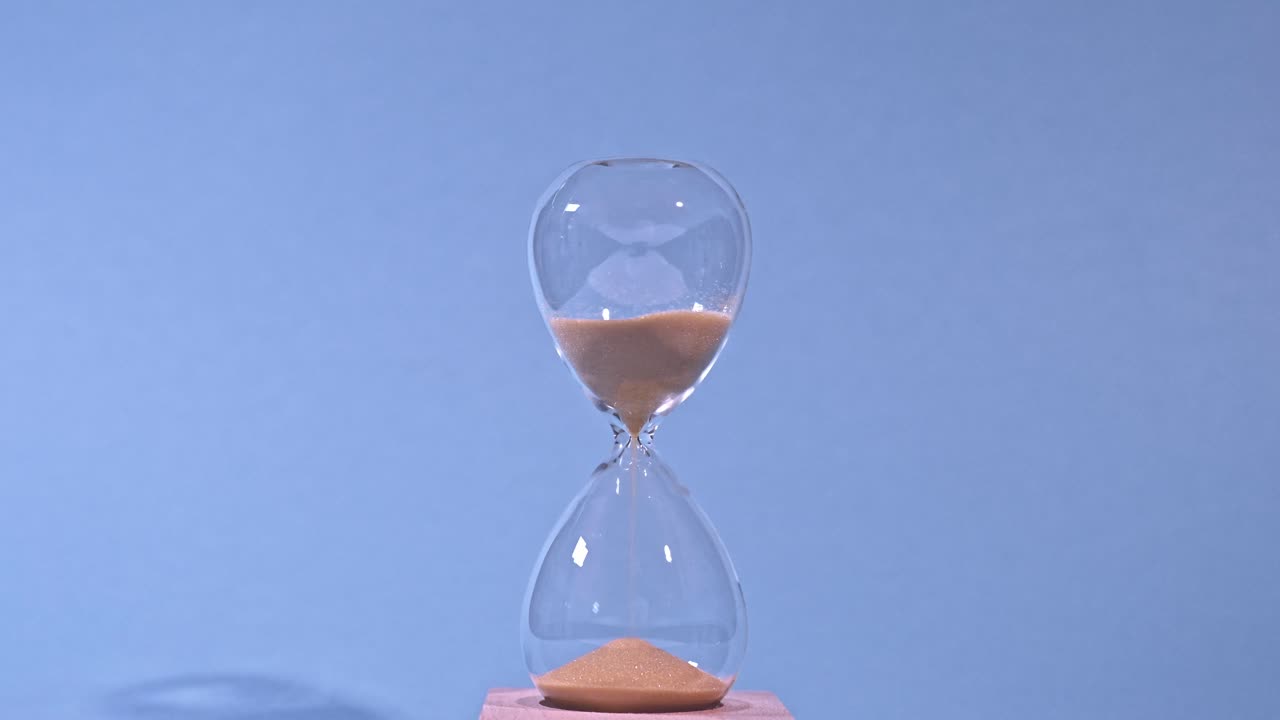 A transparent hourglass with golden sand flowing from the upper chamber to the lower one, set against a soft blue background. The video symbolizes the passage of time and the concept of countdown.