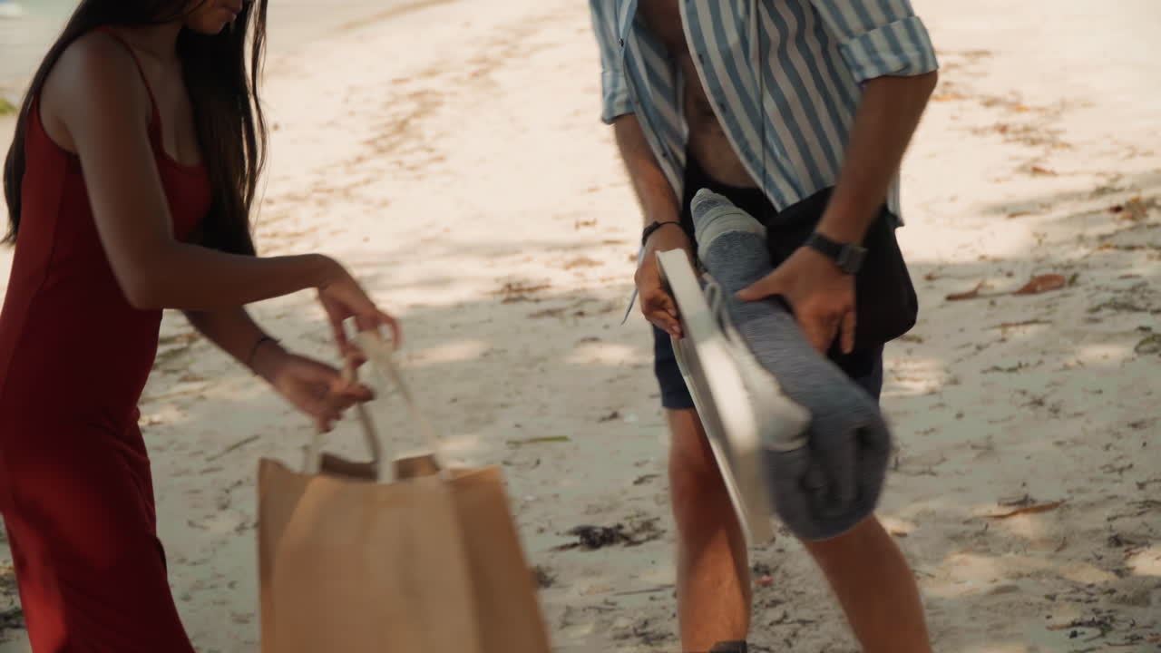 Stylish couple arriving for beach picnic white man and asian woman unpack straw bag on weathered chair with dog nearby, open shirt and red dress, sunglasses, towel, boats bobbing in sunlit cove