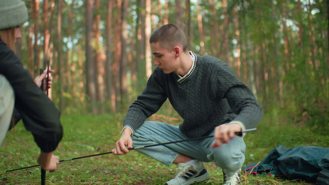 Lady hands tent pole to husband who squats and carefully fixes it during outdoor camping setup in forest surrounded by greenery and equipment as they prepare shelter together with teamwork
