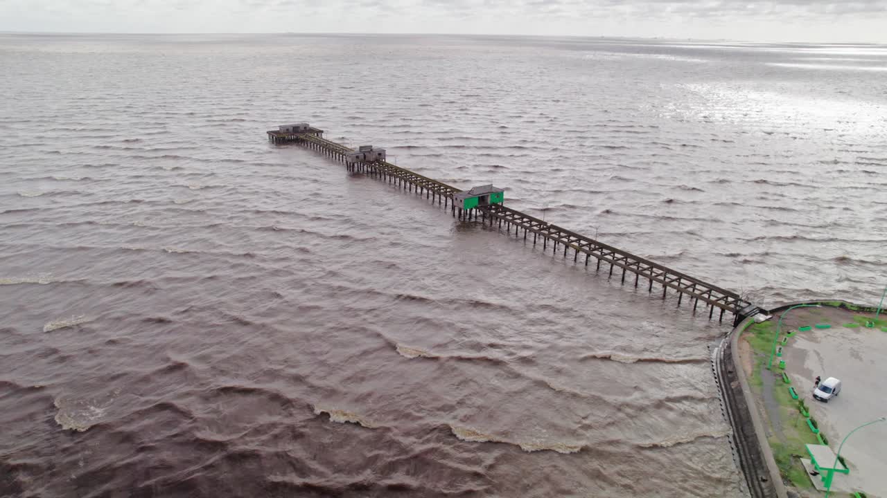 Aerial orbit shot of a pier extending into the Rio de la Plata, Punta Lara, Buenos Aires. The drone highlights the pier's structure, smooth water, and surrounding natural landscape