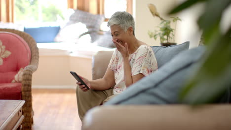 Glückliche-ältere-Frau-Mit-Gemischter-Abstammung,-Die-Auf-Der-Couch-Sitzt-Und-Zu-Hause-Ihr-Smartphone-Benutzt,-Zeitlupe