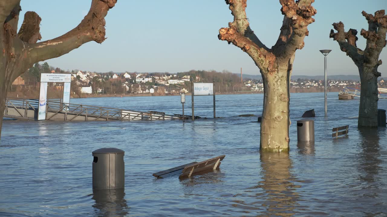 Benches, trash cans, street lamps and trees standing in the flood water with river and city in the background. Medium shot, tilting up.