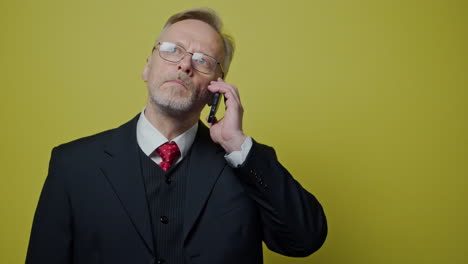 serious elderly man talking the mobile phone. senior businessman in black suit answering the call on the yellow background.