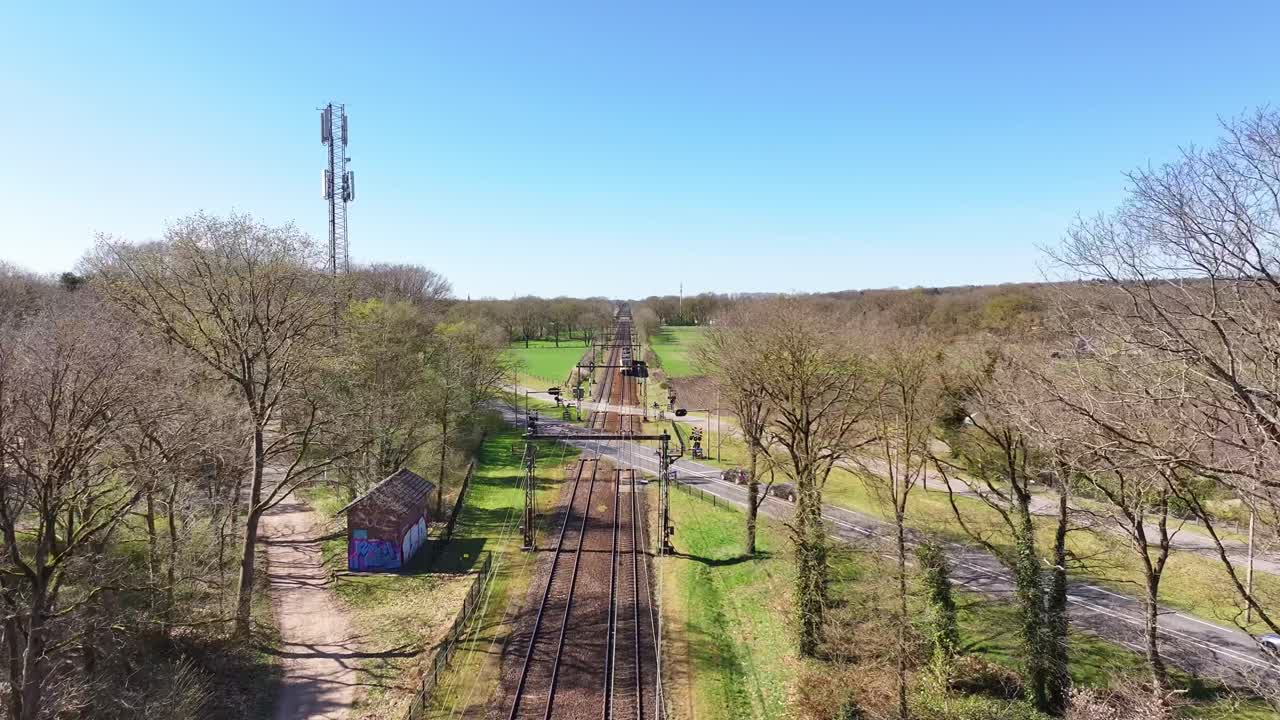 Drone follows railway from train’s view during the day, crossing a road with cars and surrounded by greenery and nature.