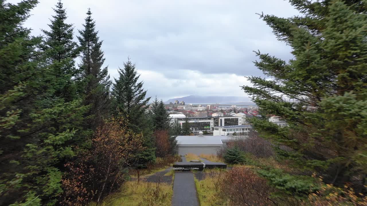 Dron volando a través de un sendero en un bosque de pinos hacia la Hallgrímskirkja de Reikiavik, en una colina, luego ascendiendo sobre los árboles para revelar Hlíðarendi y la ciudad de Reikiavik, vista aérea con dron