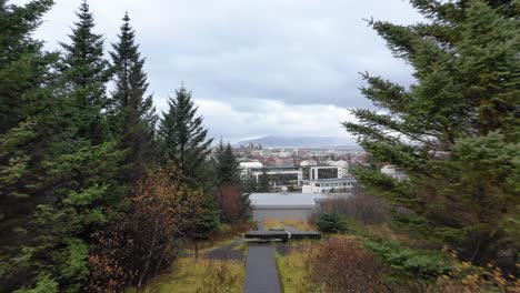 Drone-Volando-A-Través-Del-Camino-Del-Bosque-De-Pinos-A-Reykjavik-Hallgrimskirkja,-En-La-Colina,-Luego-Ascendiendo-Por-Encima-De-Los-árboles-Para-Revelar-Hlíðarendi-Y-La-Ciudad-De-Reykjavik,-Antena-Drone