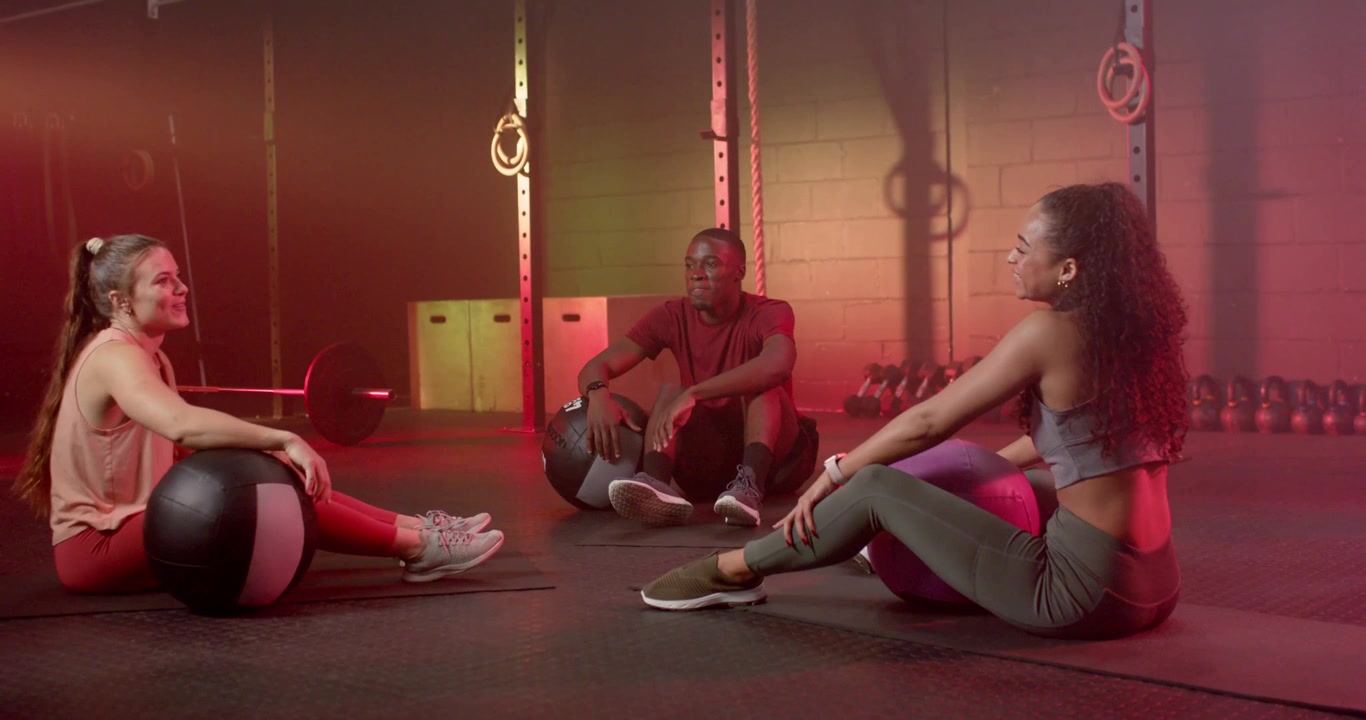 Diverse gymmates in sportswear sitting with medicine balls and stability ball, resting between sets