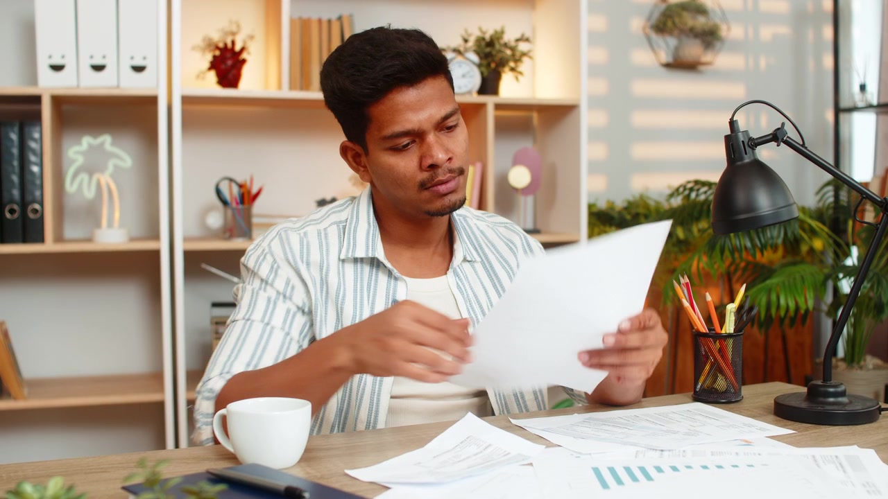 Indian man on home table drinking coffee and checking documents during focused work in living room