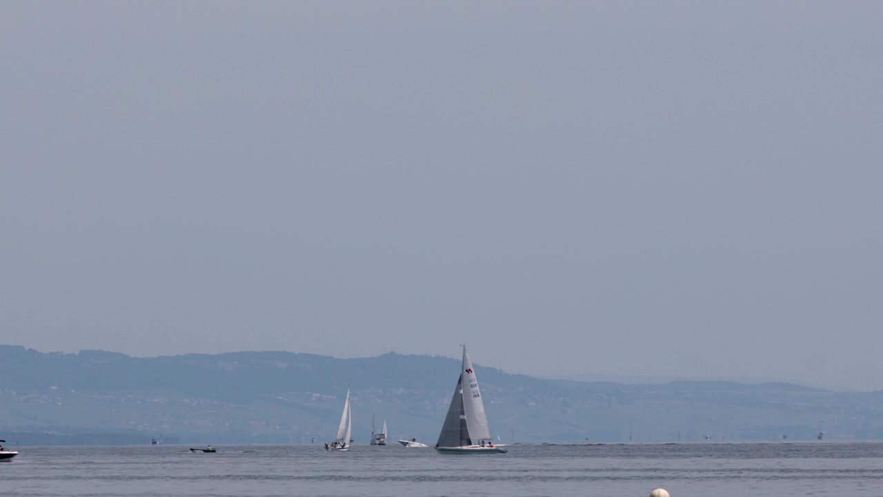 Ampia inquadratura di barche a vela che scivolano su acqua calma sotto cieli sereni con catene montuose distanti.