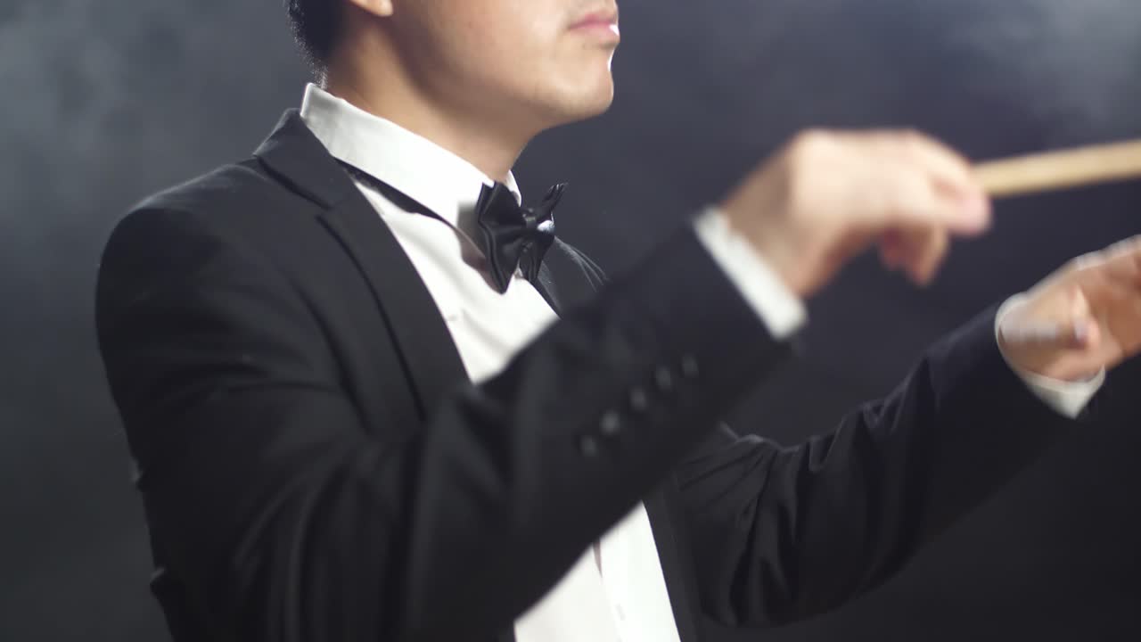 안개가 가득한 검은 스튜디오에서 눈을 감고 몸짓을 보이는 아시아계 지휘자 남자.