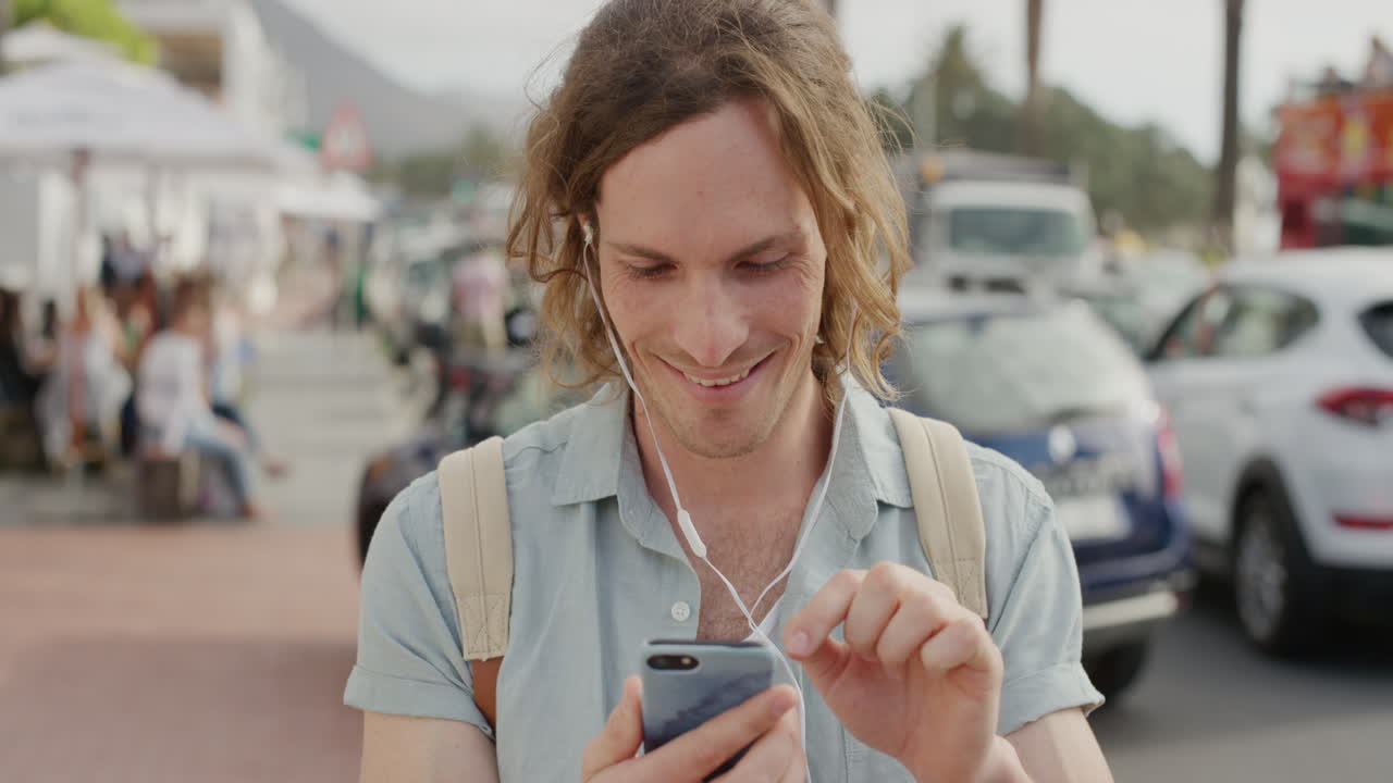portrait of young man using smartphone texting sending messages browsing wearing earphones enjoying relaxed summer day on sunny beachfront street mobile communication