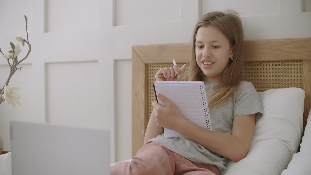 학교 아이 들 을 위한 온라인 교육 소녀 는 수업 을 위해 노트북 과 펜 을 들고 침대 에 앉아서 집 에서 학습 하고 있다