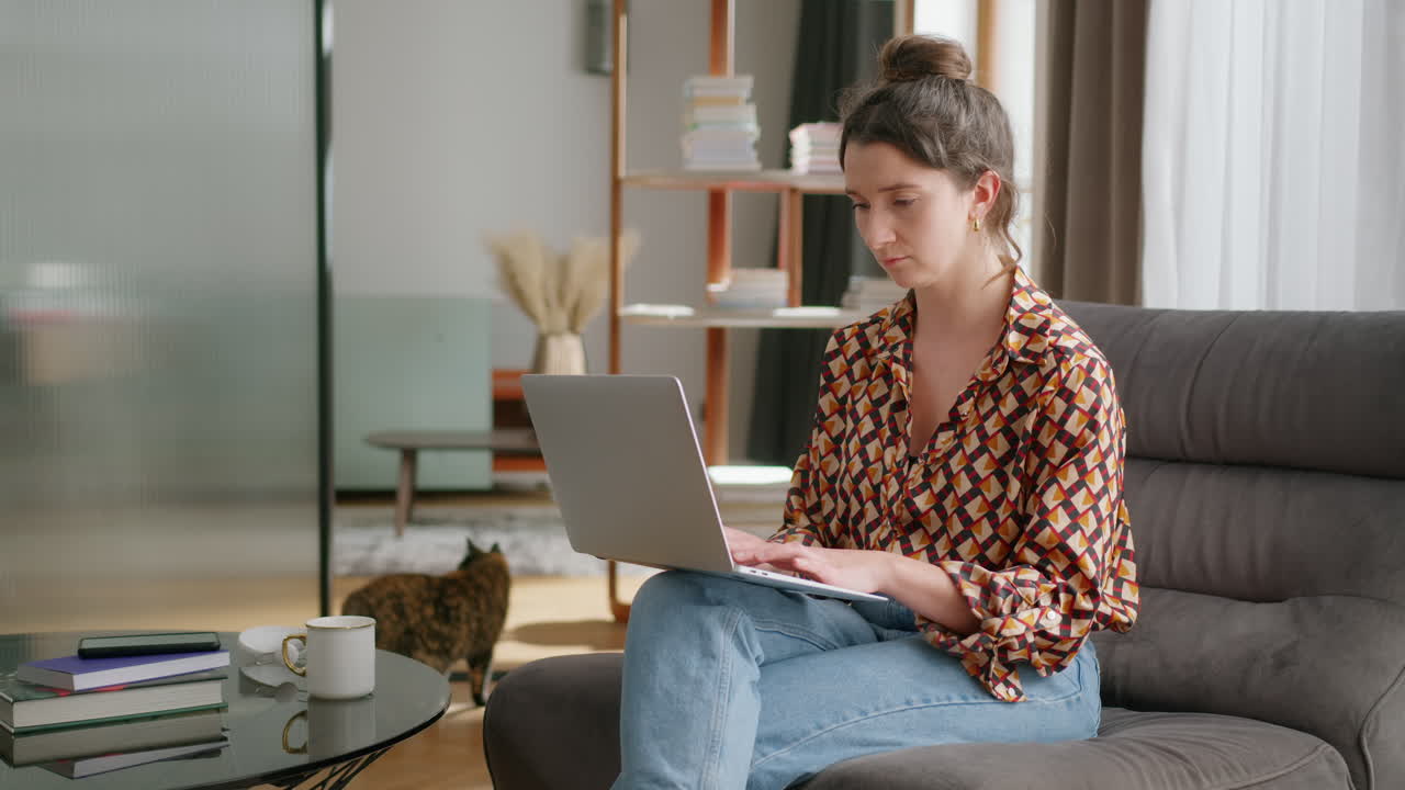 Woman uses laptop in living room while cat walks in background, slomo