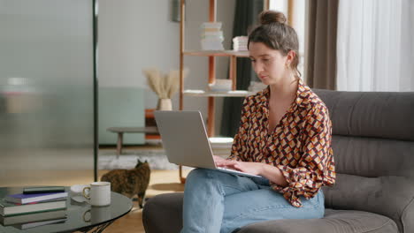 Una-Mujer-Usa-Una-Computadora-Portátil-En-La-Sala-De-Estar-Mientras-Un-Gato-Camina-En-Segundo-Plano,-En-Cámara-Lenta