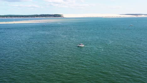 Arcachon-Bay-Francia-Con-Gente-En-Una-Lancha-Con-Duna-De-Arena-Du-Pilat-En-El-Fondo,-Dolly-Aéreo-A-La-Izquierda-Revela-Disparo