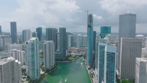 Aerial-slide-and-pan-footage-of-group-of-tall-downtown-skyscrapers.-Boats-on-river-passing-by-construction-site.-Miami,-USA