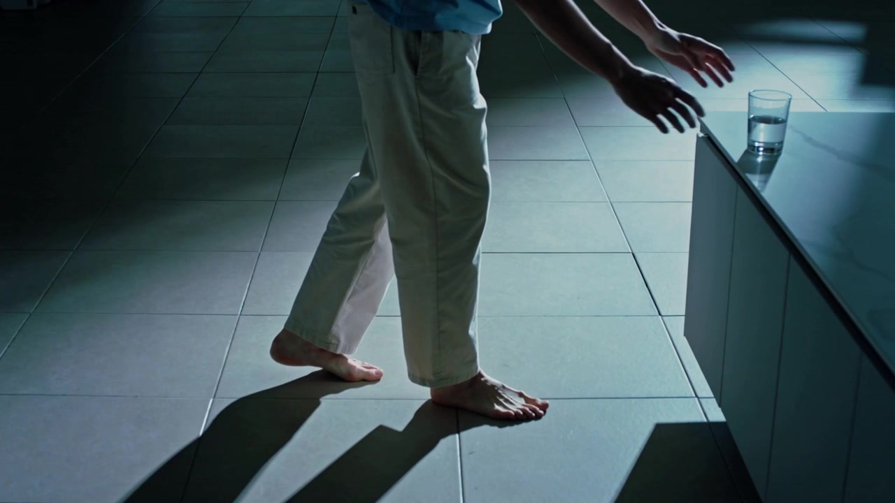 Walking barefoot shirted adult under light on kitchen tiles, reaching to pick up glass on counter