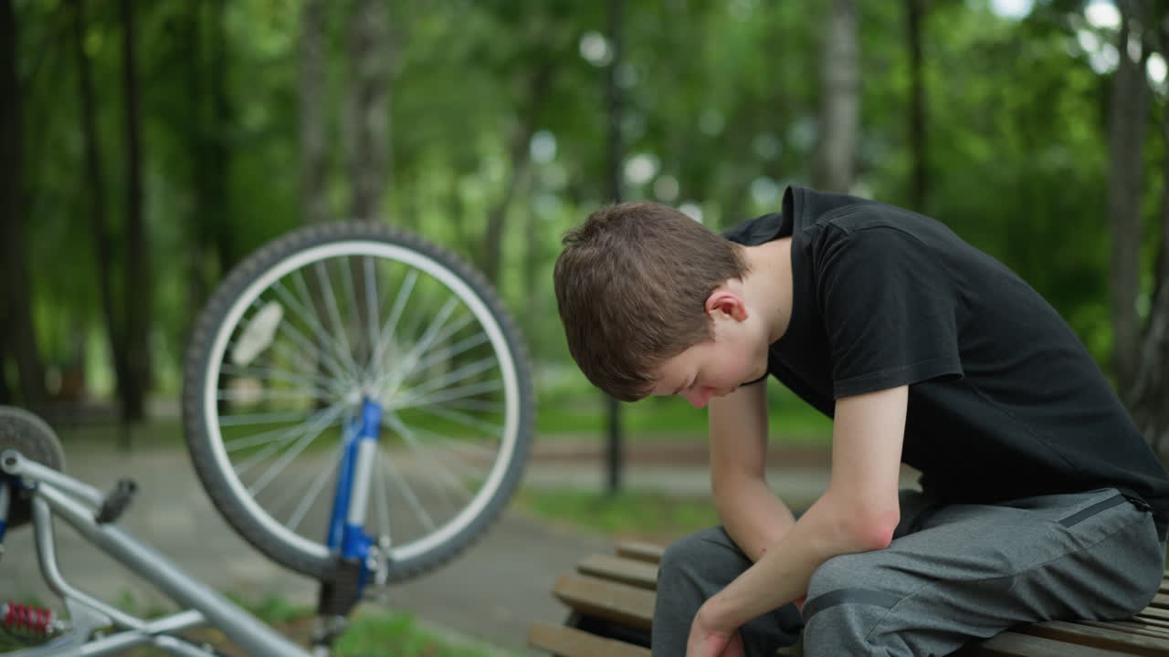 un joven con una camiseta negra se sienta con la cabeza hacia abajo, contemplando pensativo en un banco del parque, su bicicleta está al revés a su lado, con una vista borrosa de los árboles en el fondo