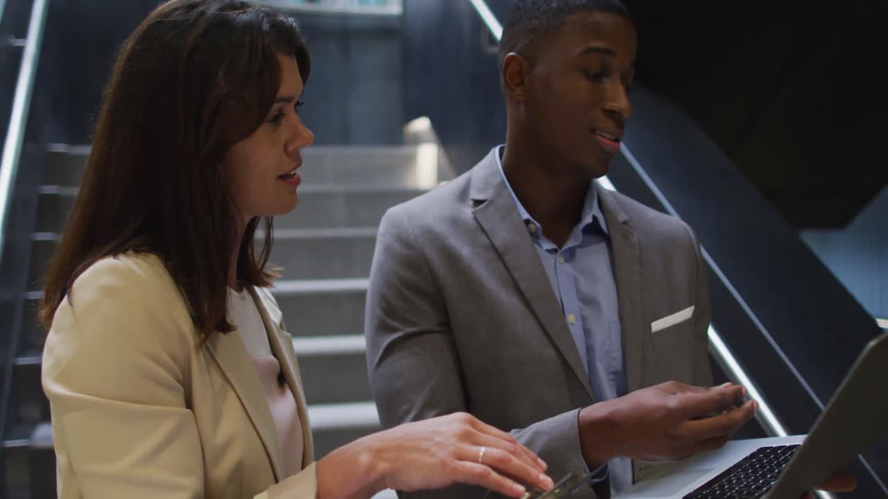 Diverse businessman and businesswoman talking and using laptop in corridor of modern office