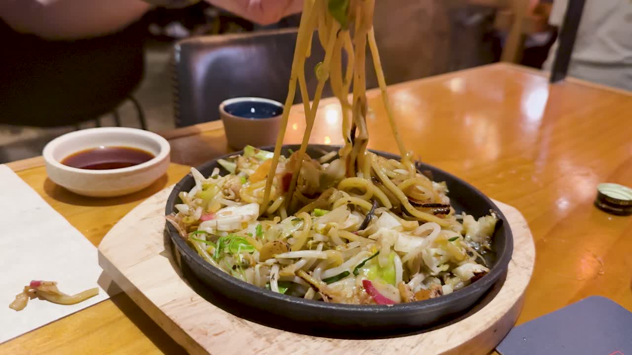 Chopsticks lift steaming noodles from a sizzling platter, surrounded by vibrant vegetables and accompanied by sake in a cozy setting
