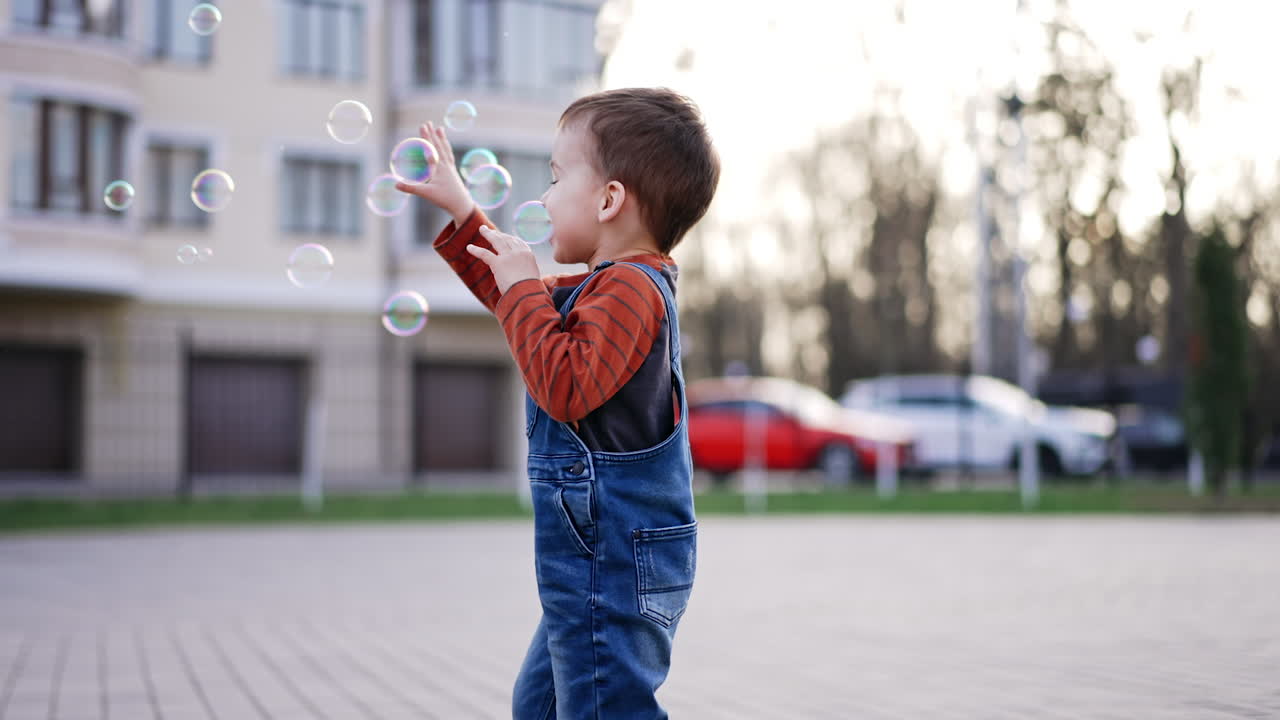 Adorable Caucasian toddler boy in jeans romper stands outdoors. Happy kid catching soap bubbles.