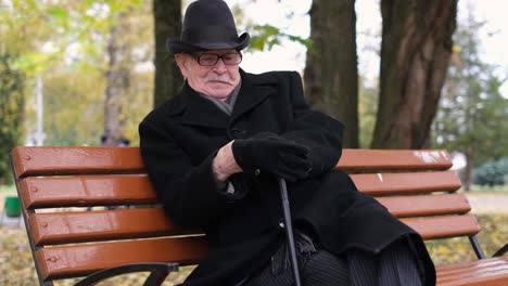 an elderly man in black clothes sits on a park bench and puts on black gloves an elderly man in black clothes sits on a park bench and puts on black gloves rest in the autumn park