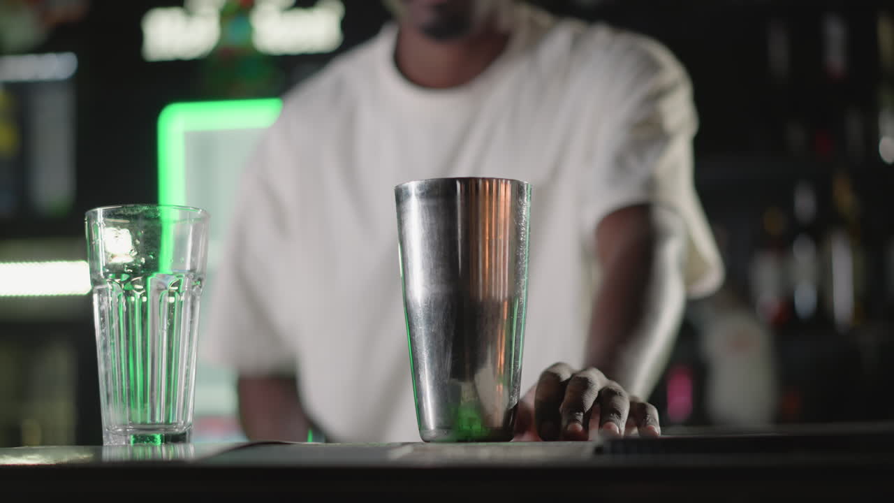 Bartender pouring ice into cocktail shaker while preparing drink at modern bar with vibrant green neon lighting, setting up for mixing, and drink creation