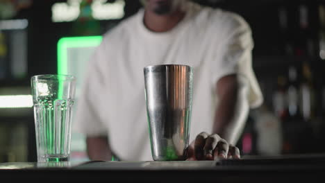 bartender pouring ice into cocktail shaker while preparing drink at modern bar with vibrant green neon lighting, setting up for mixing, and drink creation